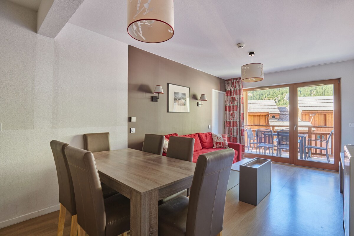 A dining area with a wooden table and chairs, a red sofa, and a view of an outdoor deck with wooden furniture.