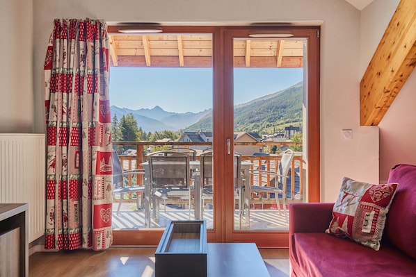 A balcony with a view of mountains and a town, a wooden table, a red sofa, and patterned curtains.