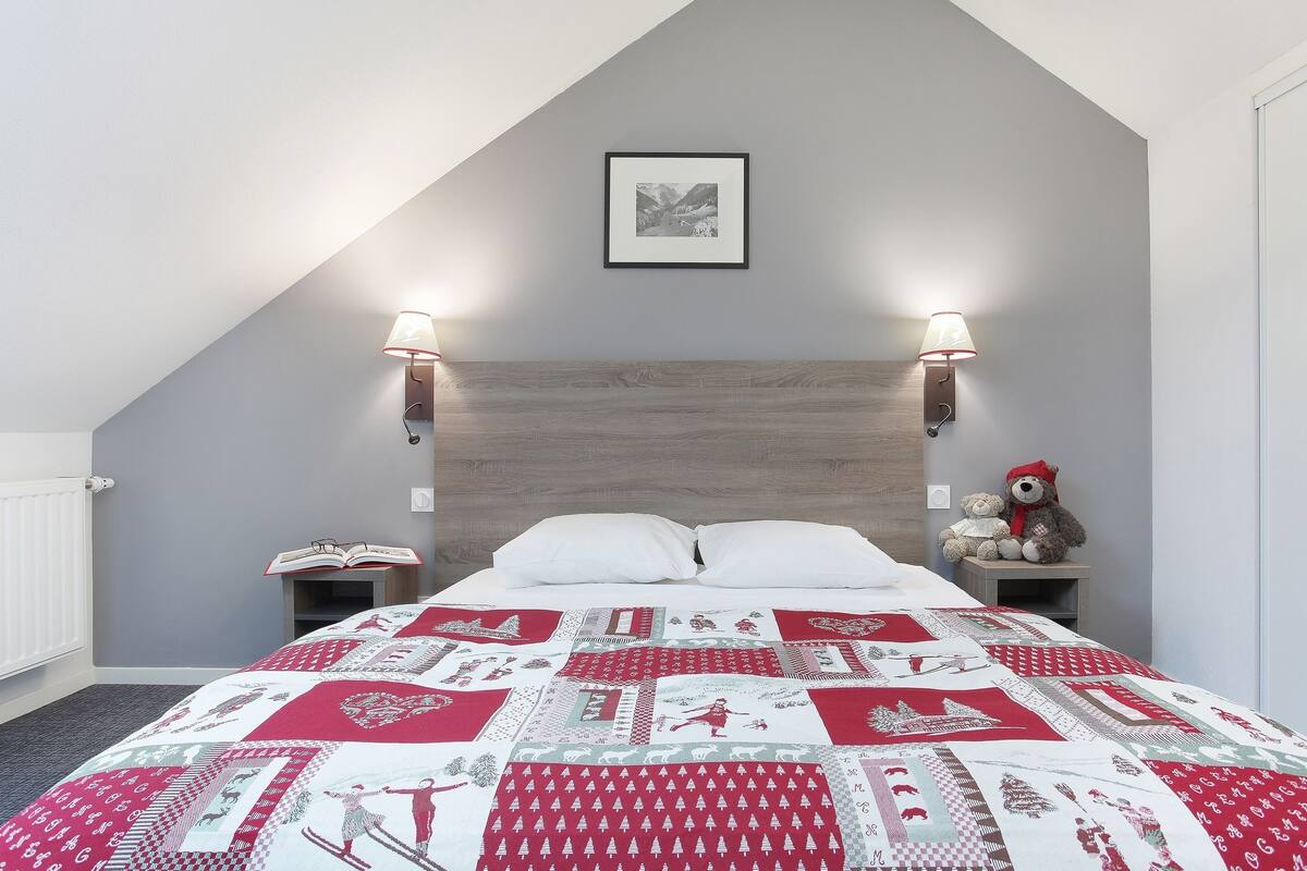 Ein Schlafzimmer mit schrägen Wänden, einer gesteppten Bettdecke, zwei Nachttischlampen, einem Teddybären und einem gerahmten Bild an der Wand.