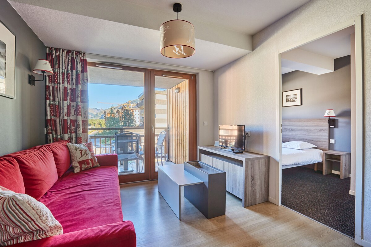 A modern hotel room with a red sofa, a desk with a flat-screen TV, a bed, and a balcony with a view of the mountains.