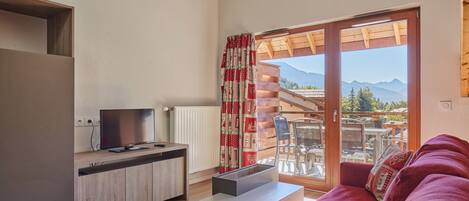 Een moderne woonkamer met een televisie, een bank en een tafel. Er is een schuifdeur die leidt naar een buitenruimte met uitzicht op bergen.