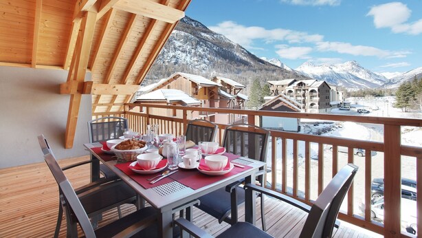 A wooden deck with a table set for dining, overlooking a mountainous landscape with snow-capped peaks.
