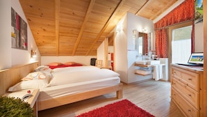 A bedroom with a wooden ceiling, a bed with white bedding, a red rug, a wooden dresser, and a bathroom with a mirror and sink. - Hotel Alegra (Livigno)