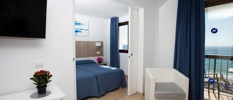 Une chambre d’hôtel avec un lit, un bureau, une télévision, un balcon donnant sur la mer et une plante en pot.