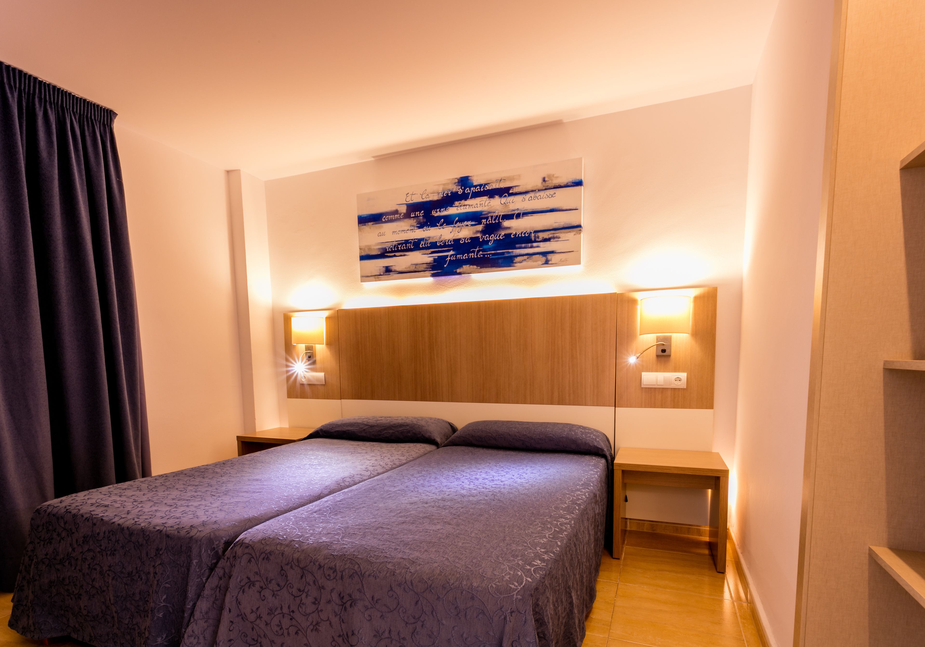 A double bed with a patterned bedspread, two bedside tables with lamps, a wall-mounted artwork, and a wooden headboard.