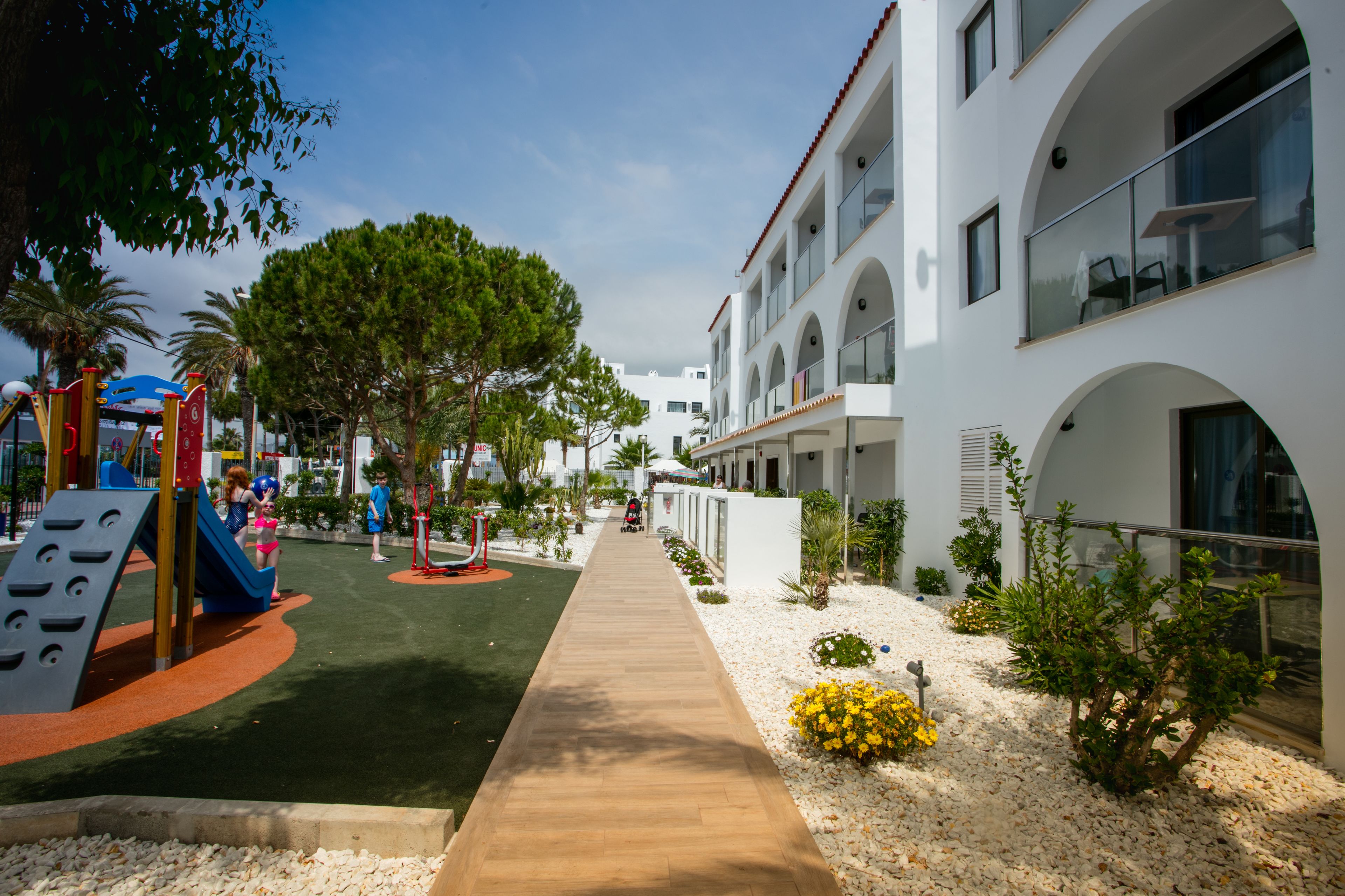 Un sendero conduce a un parque infantil con un tobogán y estructuras para escalar, rodeado de árboles y un edificio con ventanas arqueadas y balcones.