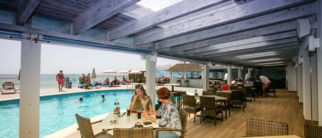 A poolside area with dining tables and chairs, overlooking a pool and beach.