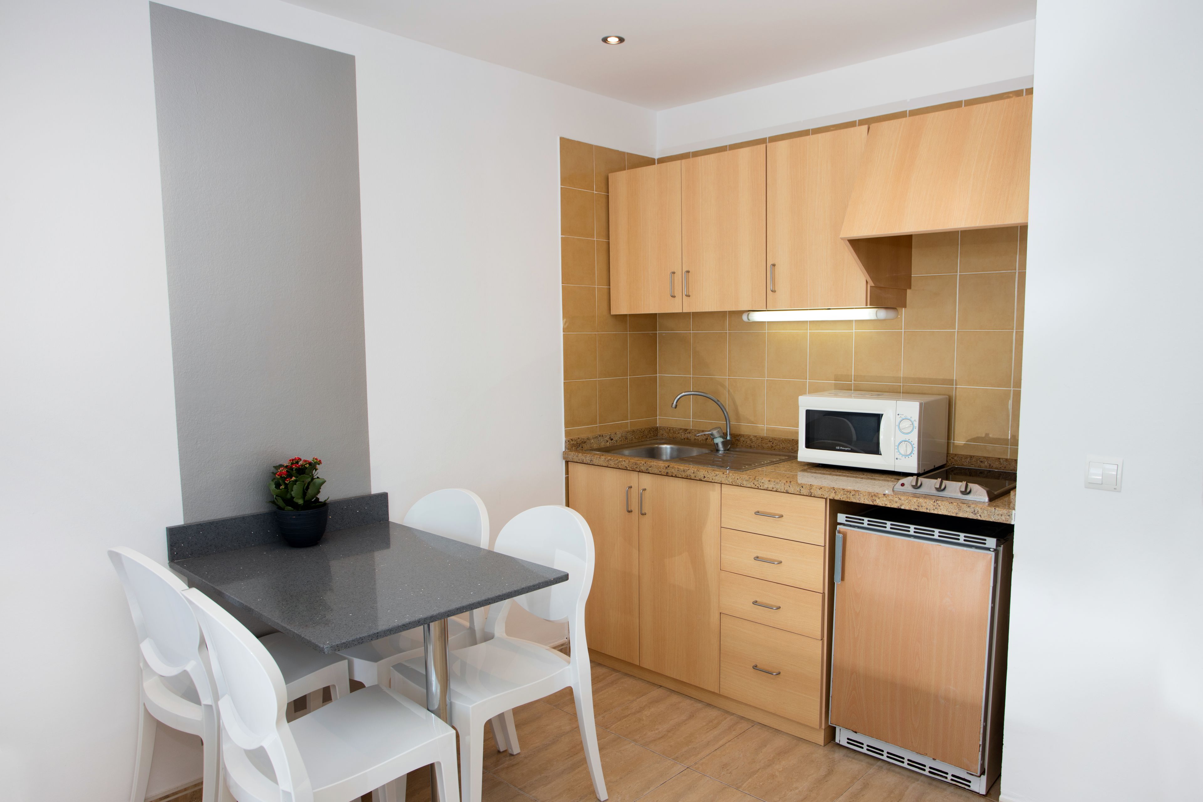 A compact kitchen with a microwave, sink, and wooden cabinets. There is a small dining table with chairs and a potted plant.