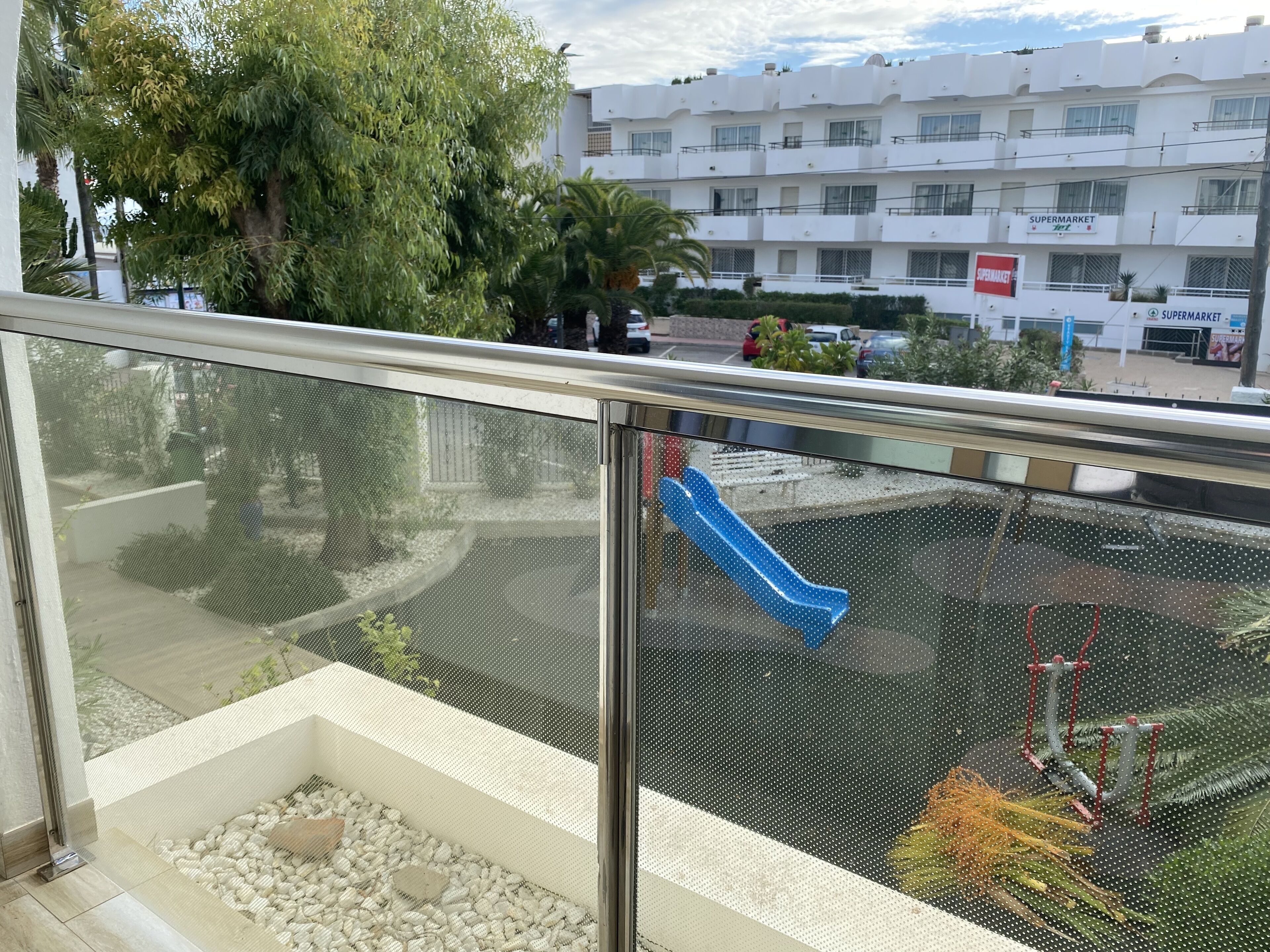 Un balcon donnant sur un terrain de jeux, un bâtiment blanc avec une enseigne de supermarché et un palmier.