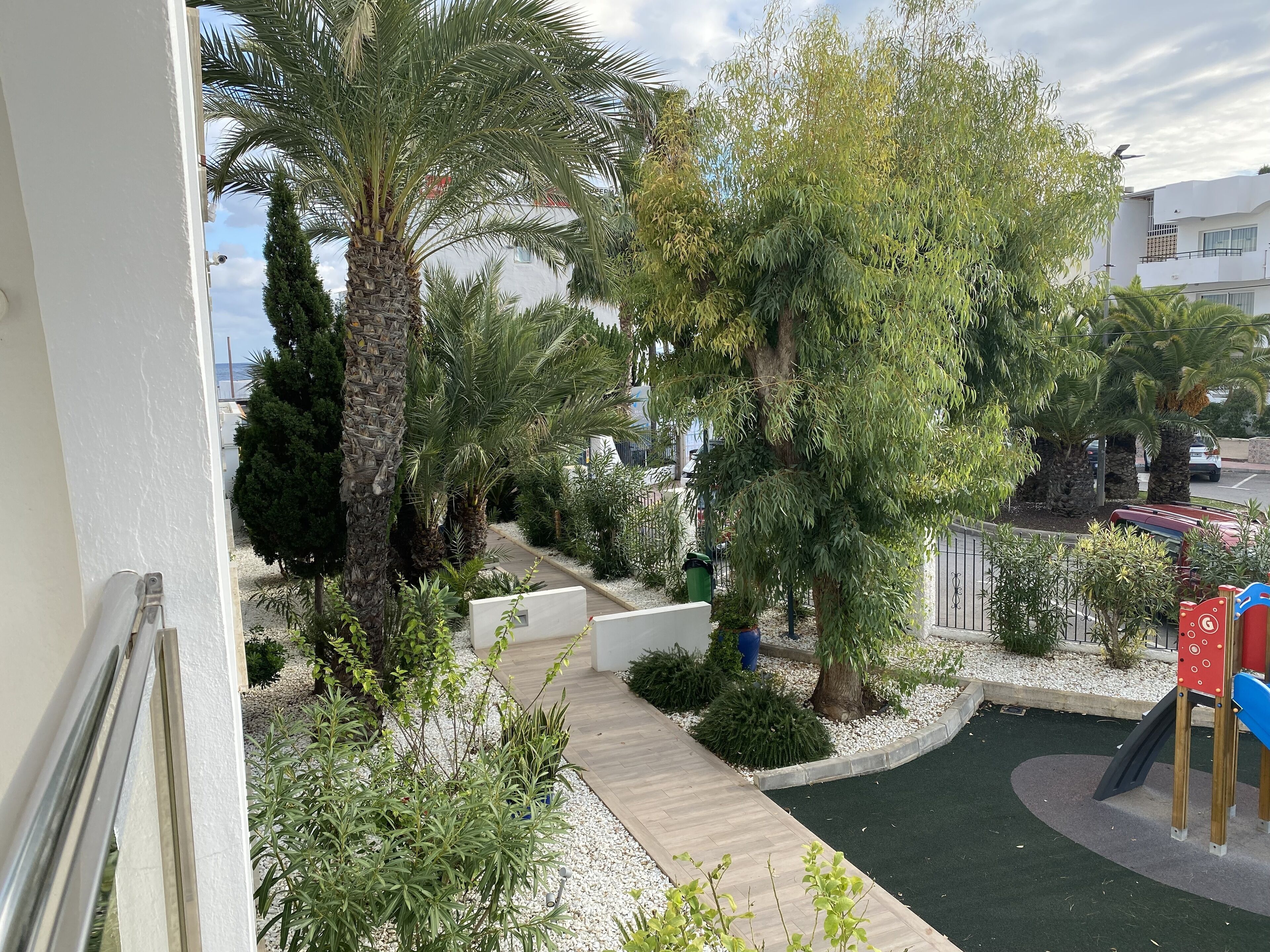 Un balcón con vista a un jardín ajardinado, que incluye palmeras, un parque infantil y un sendero.