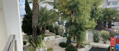 Un balcón con vista a un jardín ajardinado, que incluye palmeras, un parque infantil y un sendero.