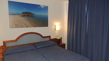 Habitación de hotel con cama, mesita de noche con lámpara, cuadro de una playa y ventana con cortinas.