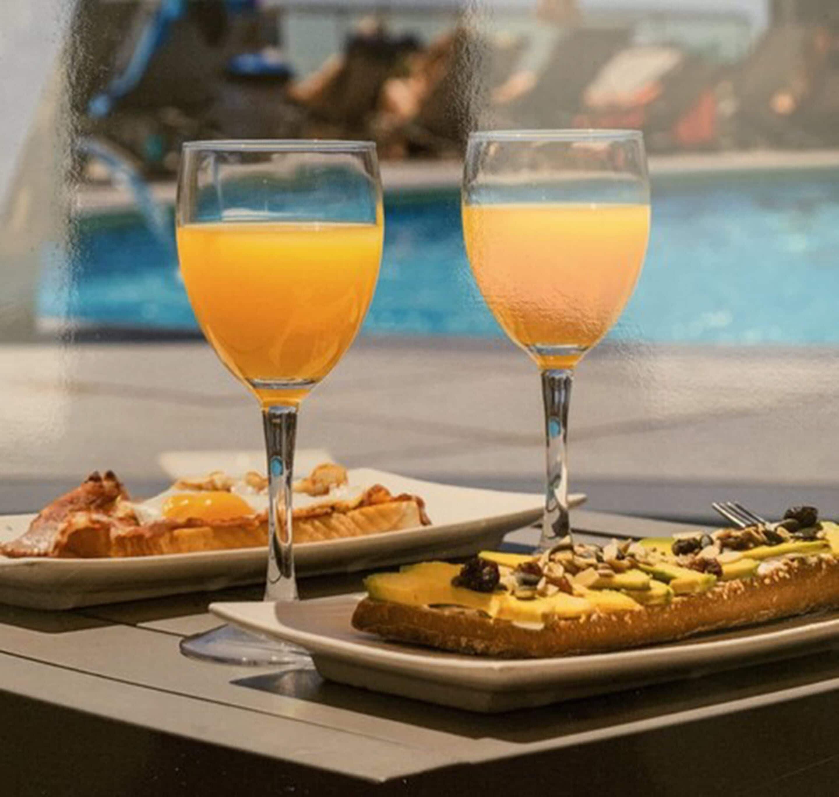 Dos vasos de jugo de naranja y un plato con alimentos para el desayuno, como tostadas y huevos, junto a una piscina.
