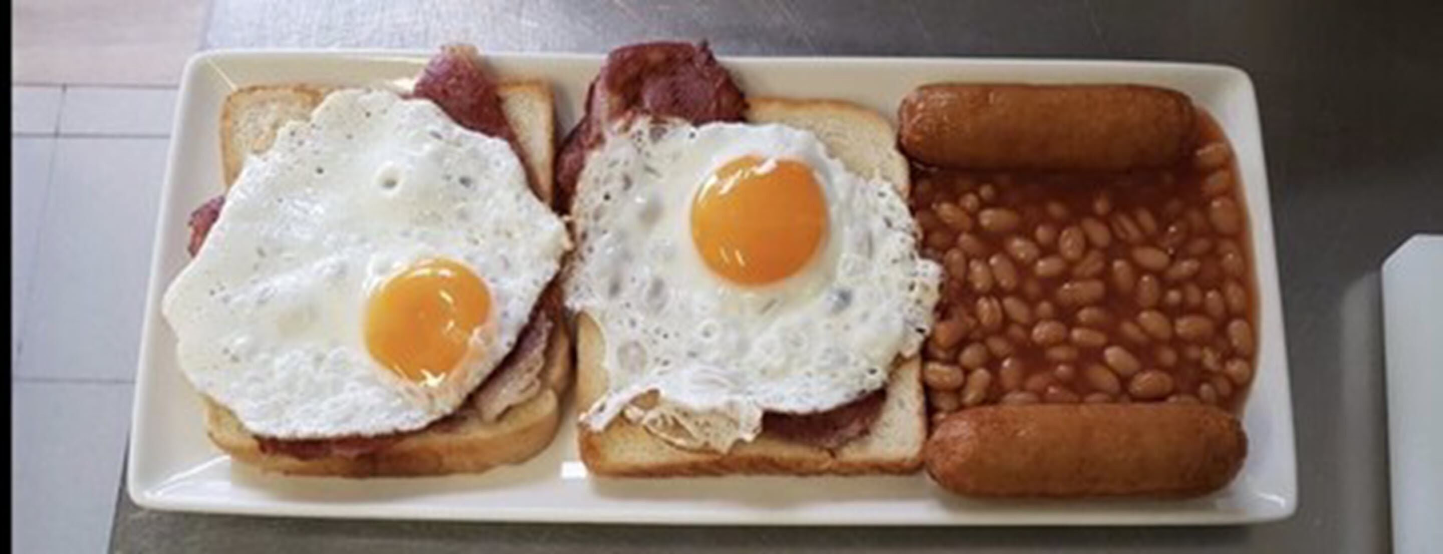 Ein Frühstücksteller mit zwei Spiegeleiern, Speck, Toast, Bohnen und einer Bratwurst.