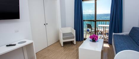 A modern hotel room with a balcony, a TV, a white desk, a blue sofa, and a view of the sea.