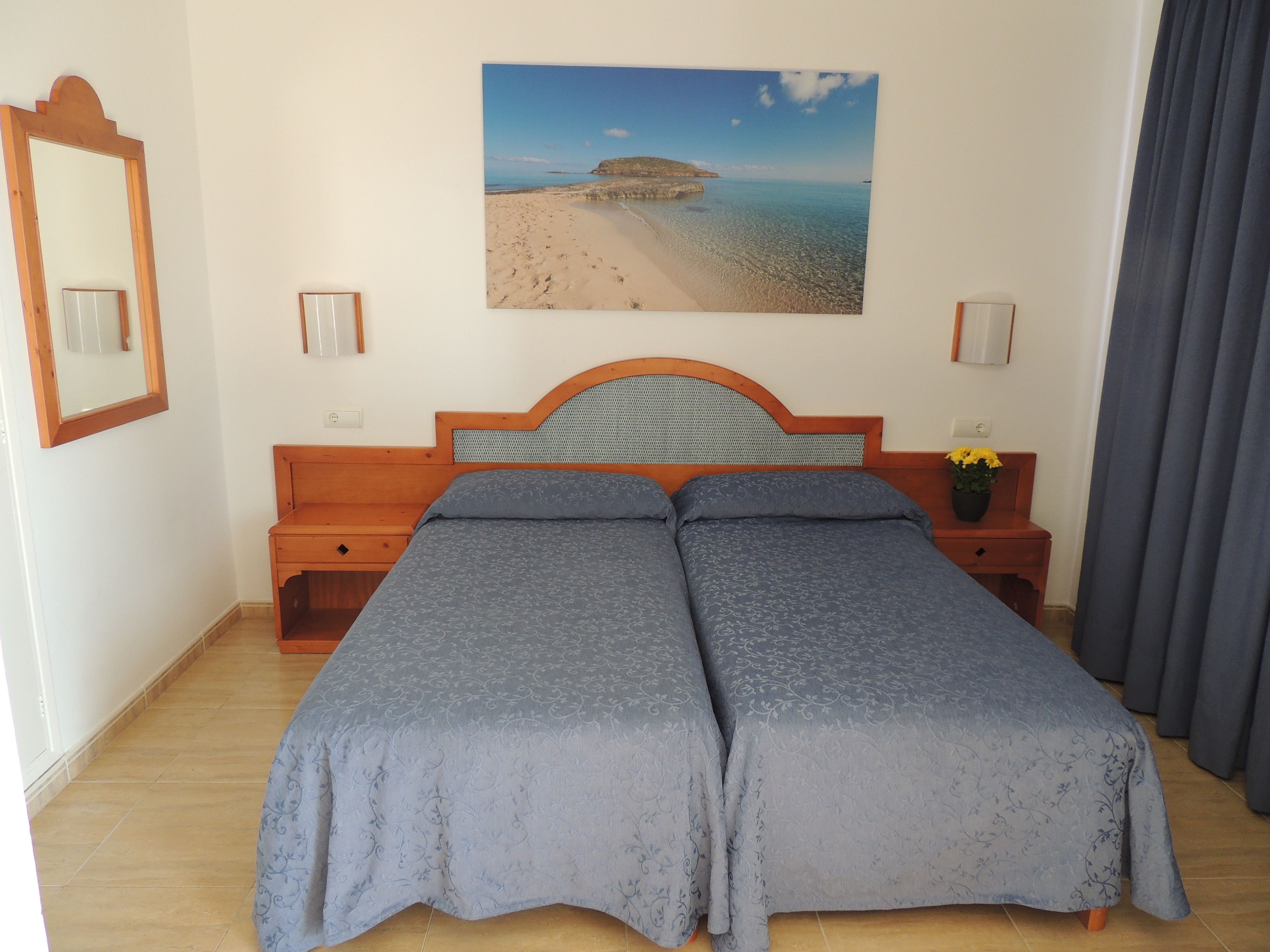 A double bed with a headboard, a nightstand with a flower vase, a painting of a beach, and a mirror on the wall.