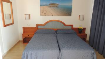 Un lit double avec une tête de lit, une table de chevet ornée d’un vase de fleurs, un tableau représentant une plage et un miroir accroché au mur.