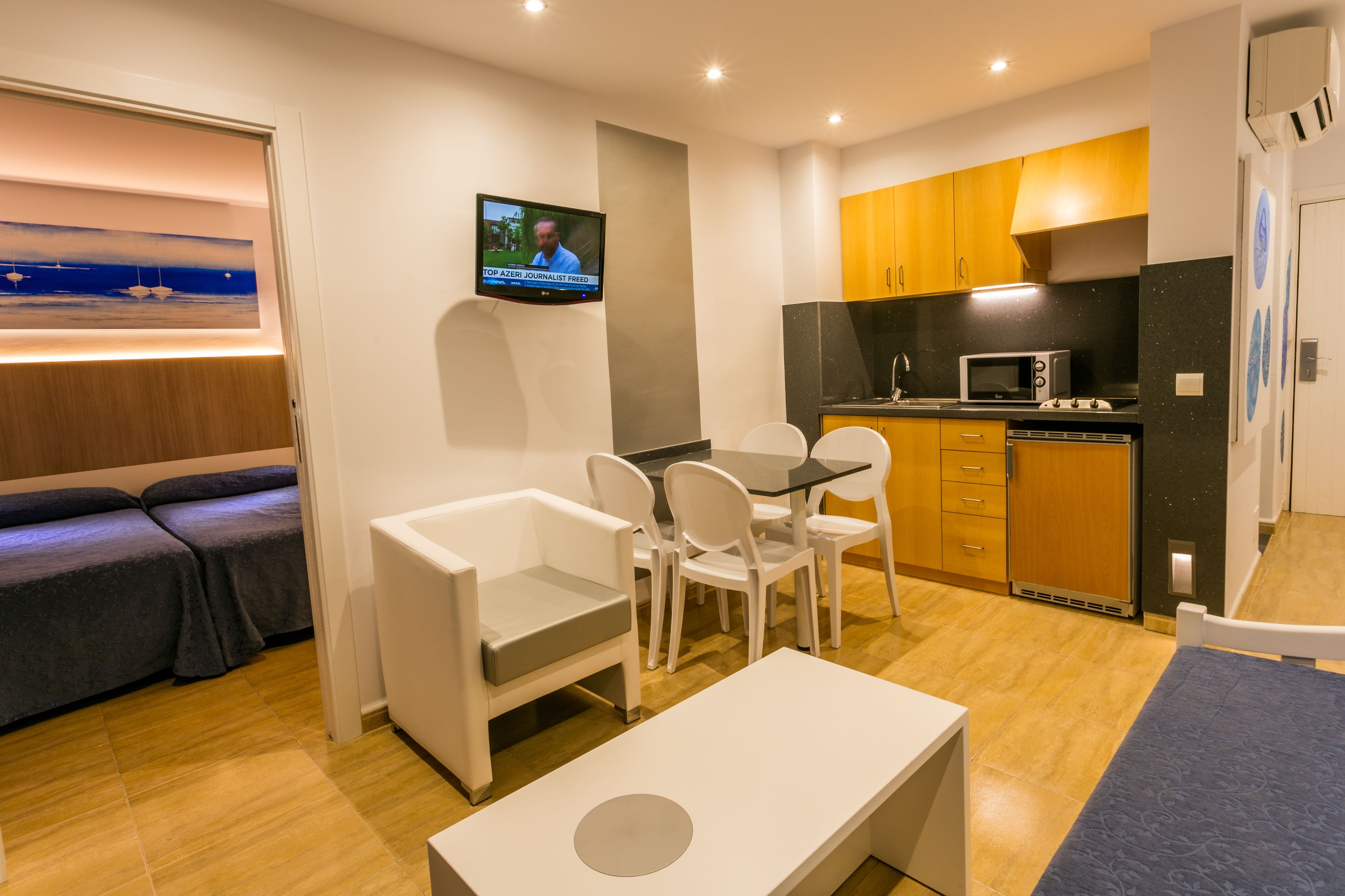 A compact living space with a sofa, a dining table with chairs, a kitchen area with a microwave, and a TV displaying a news program.