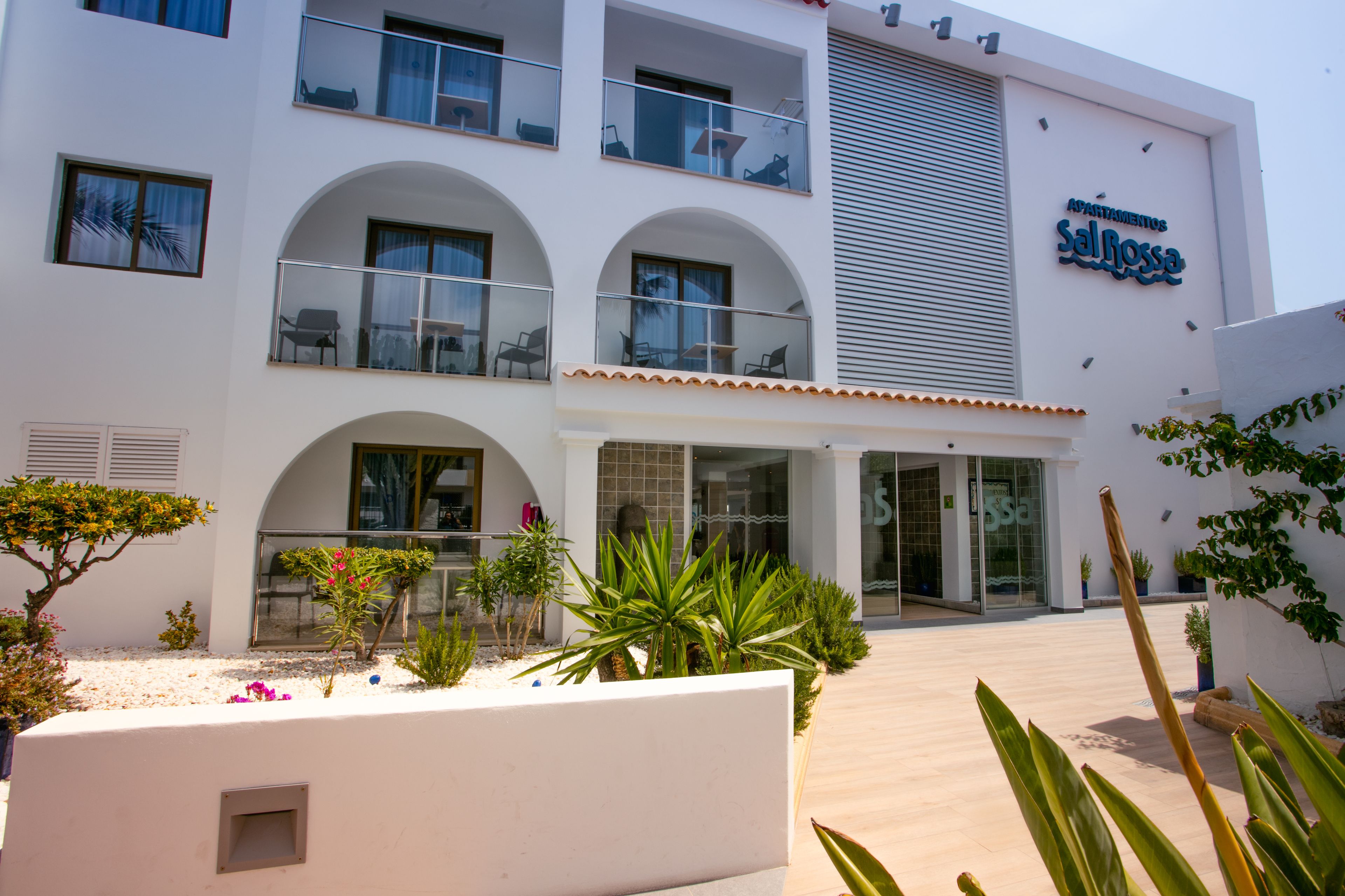 Un hotel moderno con múltiples balcones, una entrada central y un jardín bien cuidado.
