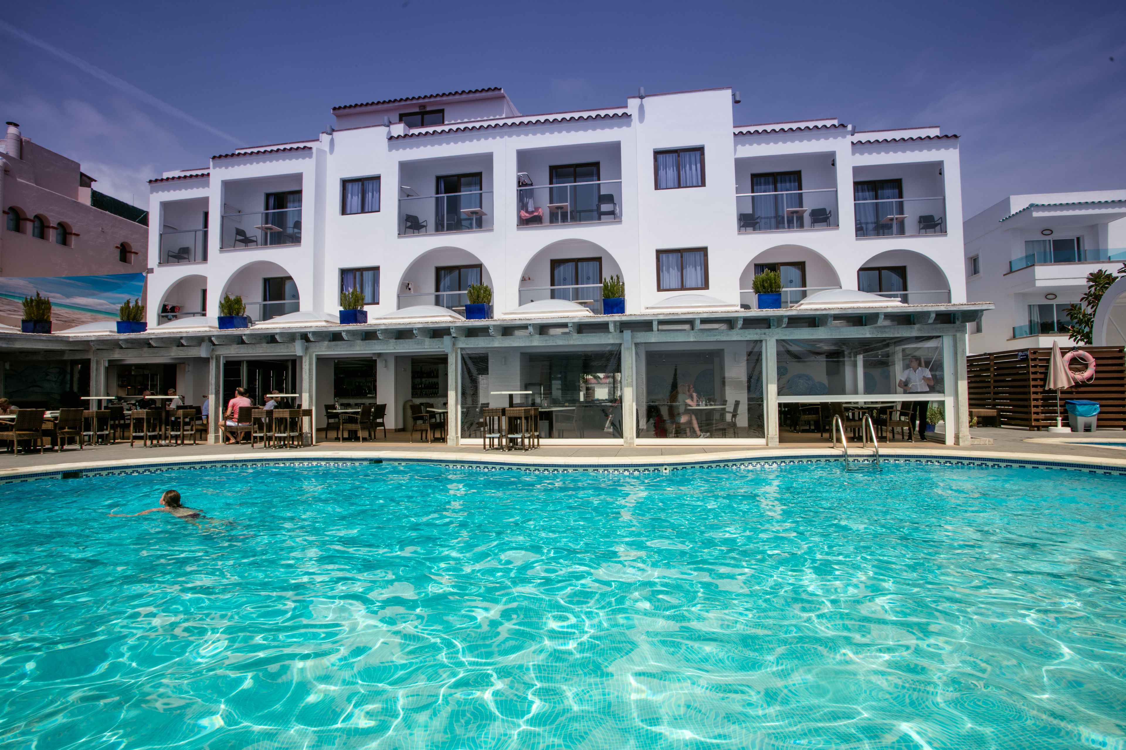 Ein Schwimmbecken mit einer Person, die schwimmt, umgeben von einem Gebäude mit mehreren Balkonen und Außenbereich-Sitzgelegenheiten.