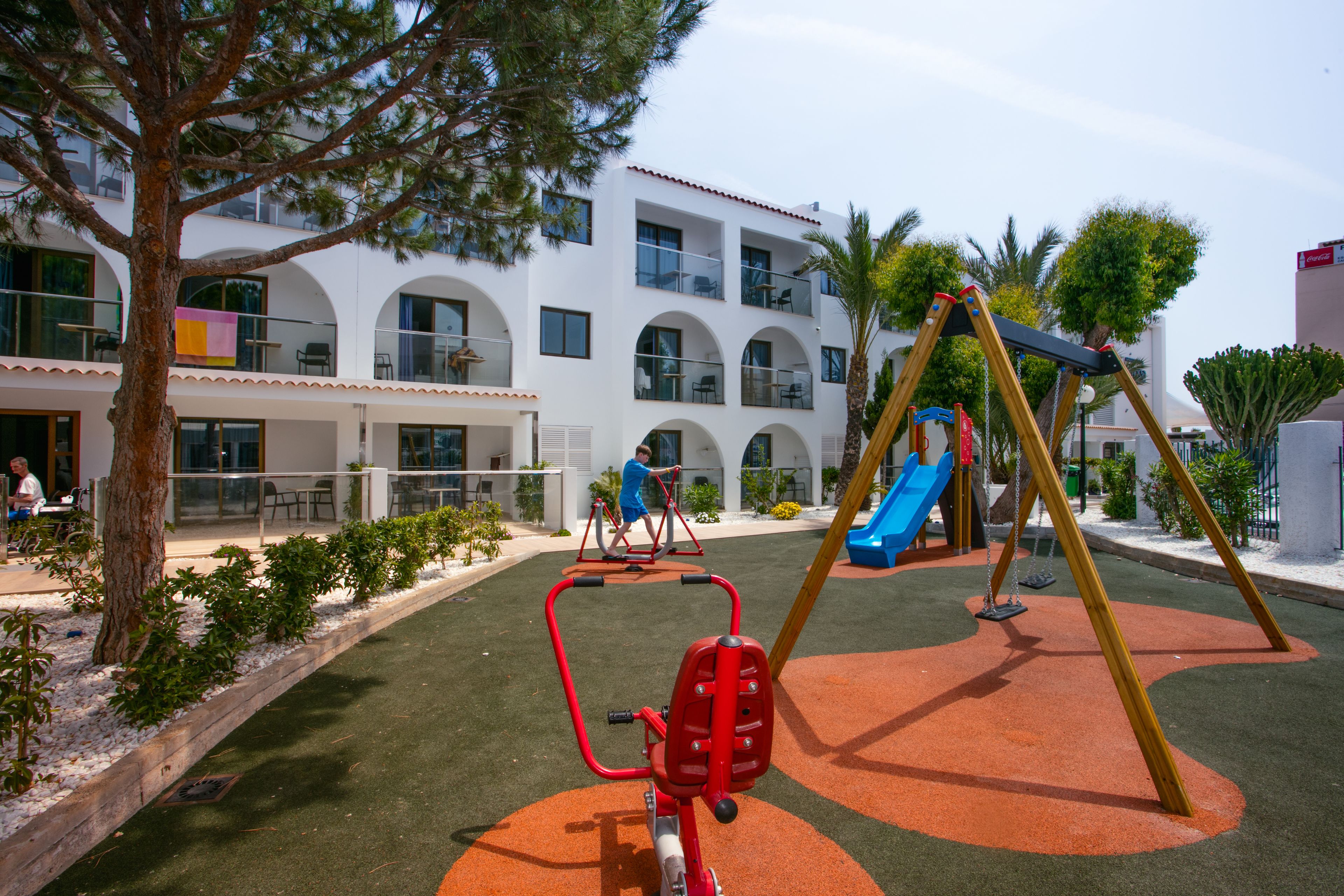 Un parque infantil con columpios y un tobogán, rodeado por un edificio blanco con ventanas de arco y balcones.