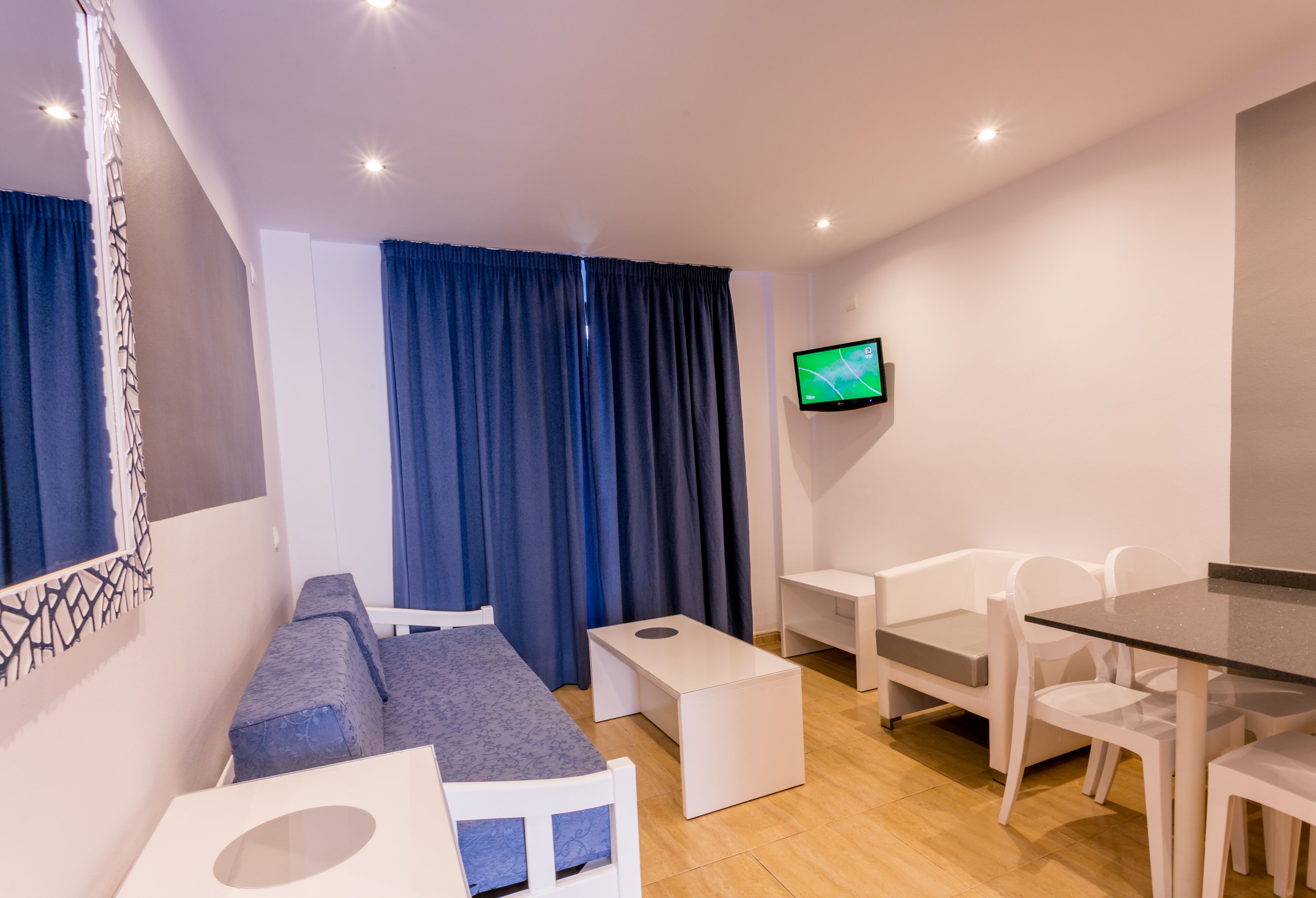 A modern room with a sofa, a small table, a glass dining table, white chairs, a TV on the wall, and blue curtains.