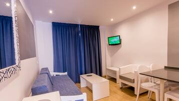 A modern room with a sofa, a small table, a glass dining table, white chairs, a TV on the wall, and blue curtains.