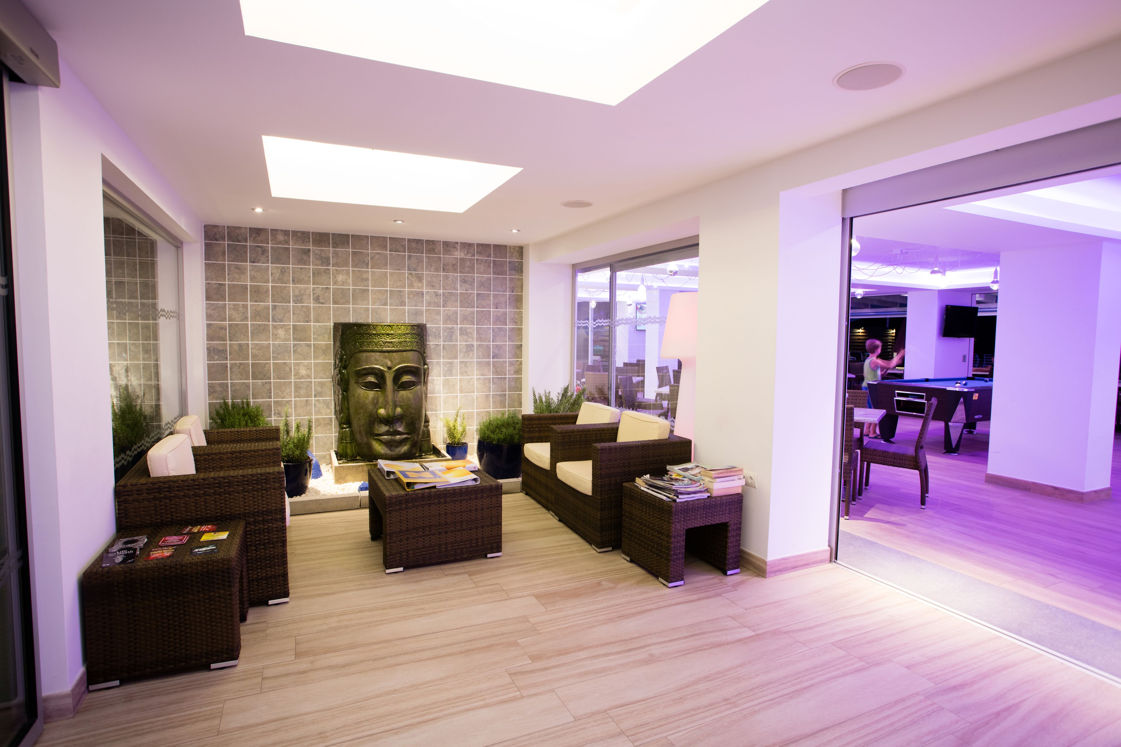 A modern lounge area with wicker furniture, a large wall-mounted sculpture, and a glass door leading to a pool table room.