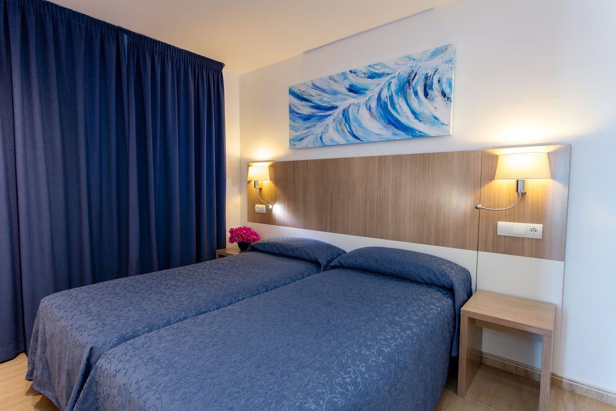 A hotel room with two beds, a wooden headboard, wall-mounted lamps, a painting, and a nightstand with a flower.