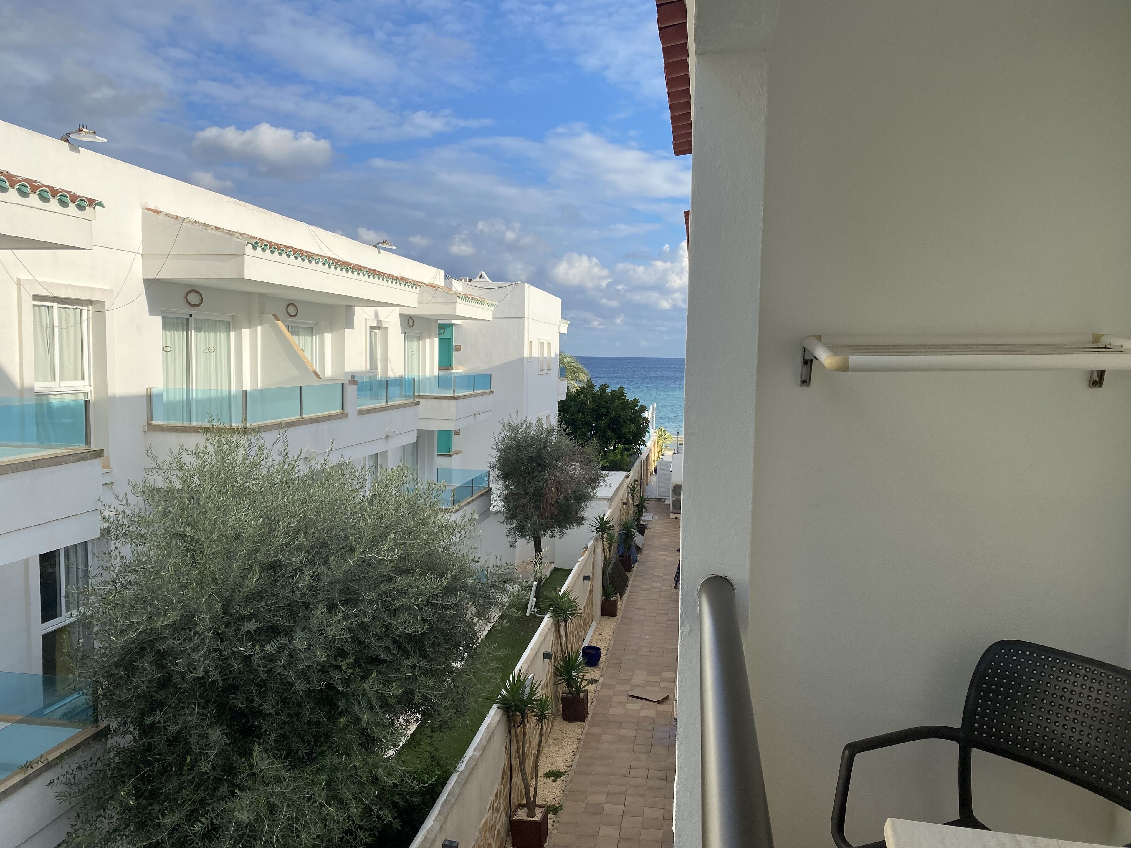 A balcony with a view of a white building, a potted plant, and a chair.