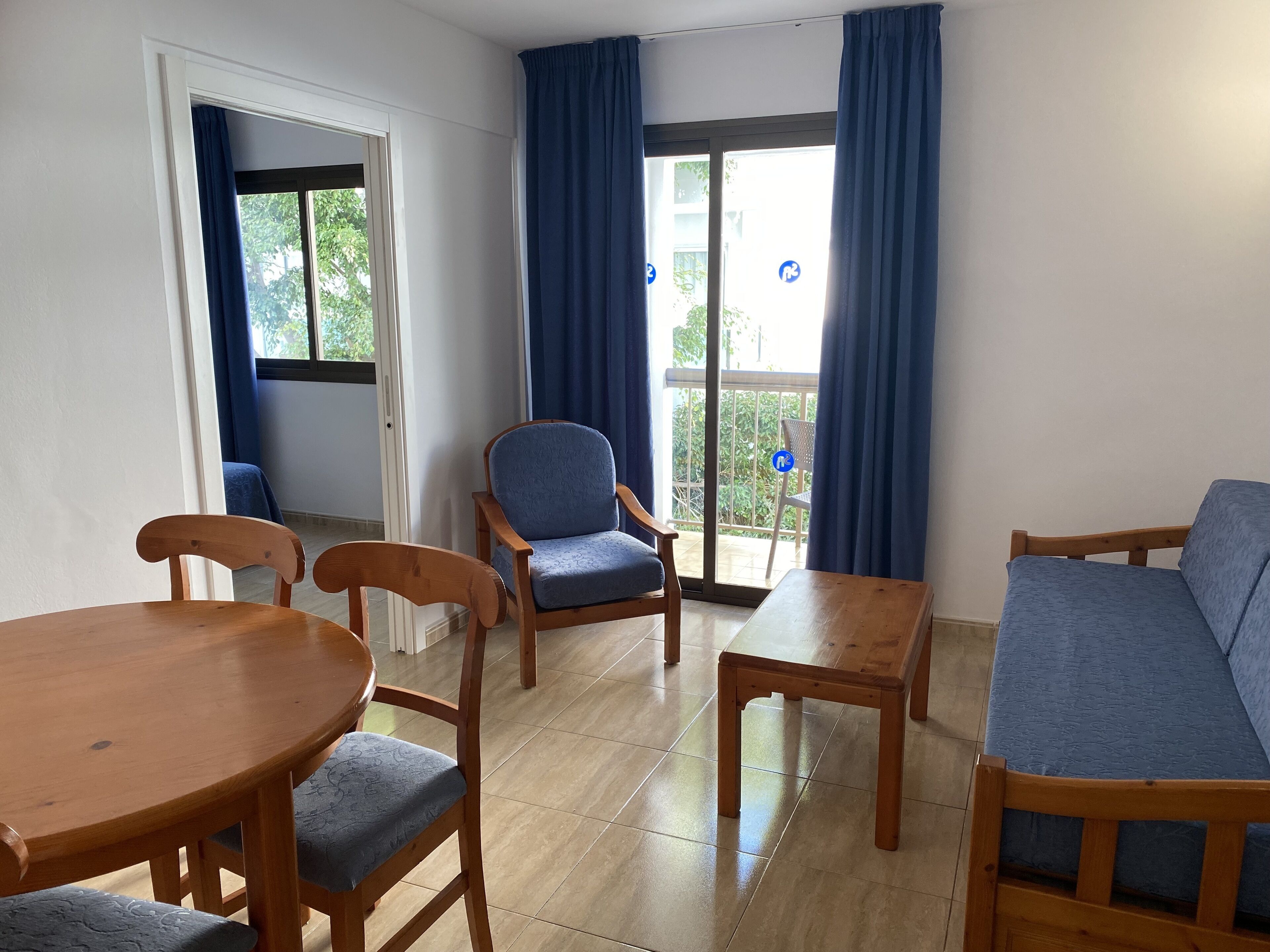 Una habitación con una mesa de madera para comer, dos sillas, un sofá y un sillón azul. Cuenta con una puerta corrediza de vidrio que da a un balcón exterior con vistas a zonas verdes.