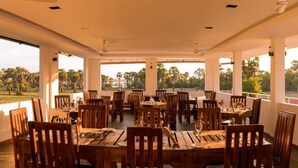 A restaurant with wooden tables and chairs, large windows with a view of palm trees and a body of water. - Palm Resort Nilaveli (Nilaveli)