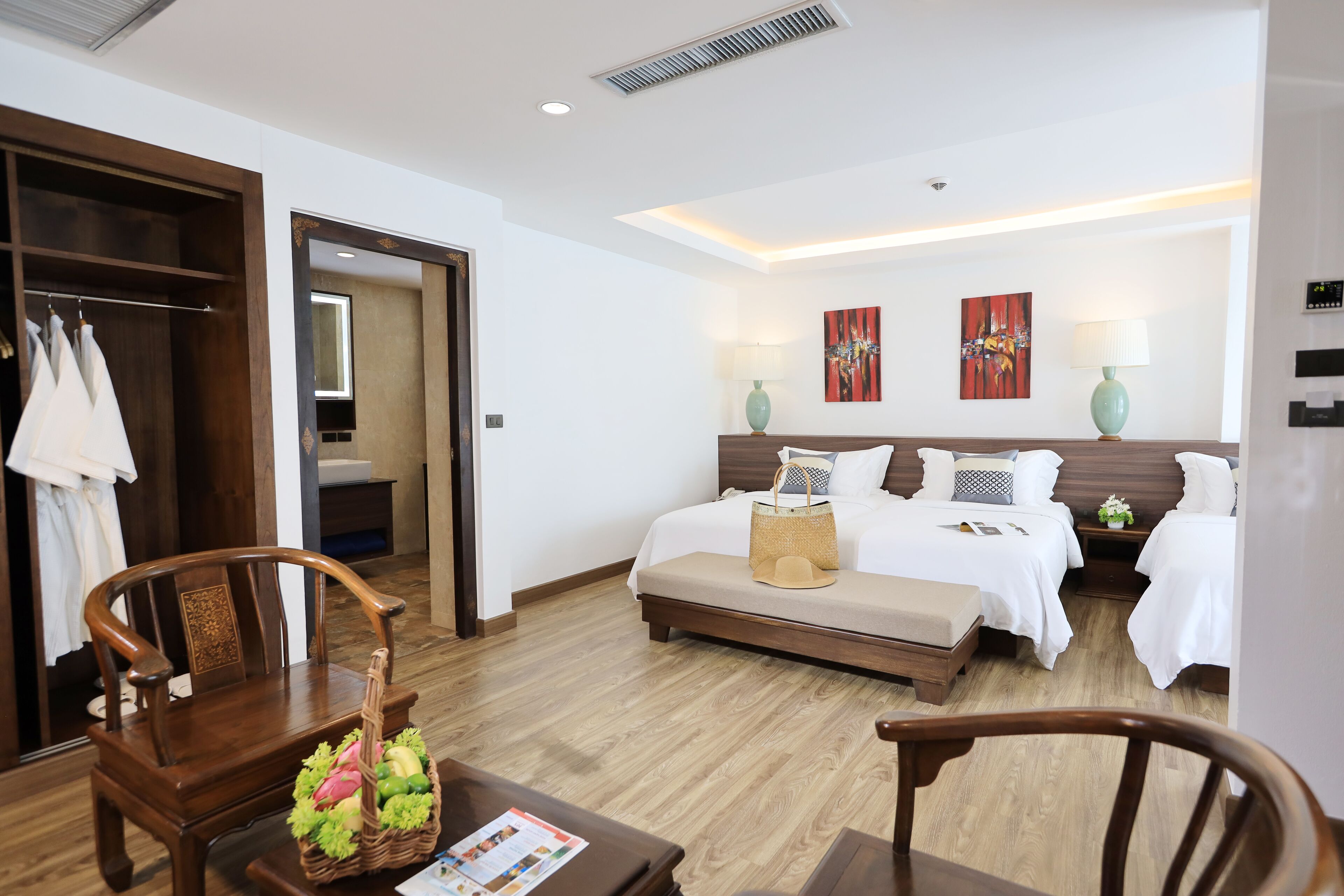 Habitación de hotel con piso de madera, una cama grande con ropa blanca, dos mesitas de noche con lámparas, una silla de madera, una pequeña mesa con una cesta de frutas y un armario con ropa colgada.