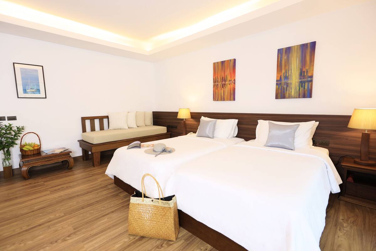 A hotel room with a large bed, two bedside tables, a nightstand, a wooden bench, a basket of fruit, and a wall-mounted painting.