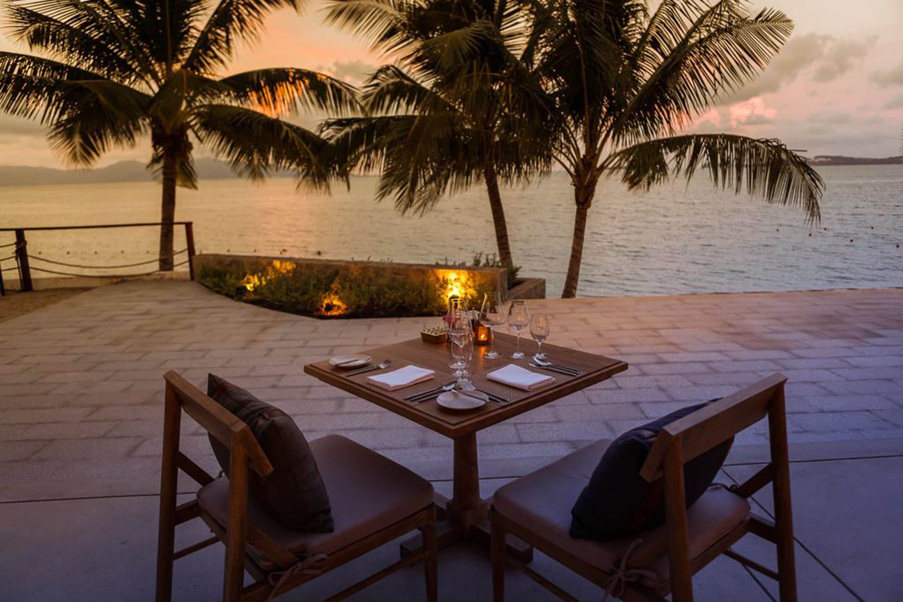 A table set for two with a view of the sea and palm trees at sunset.