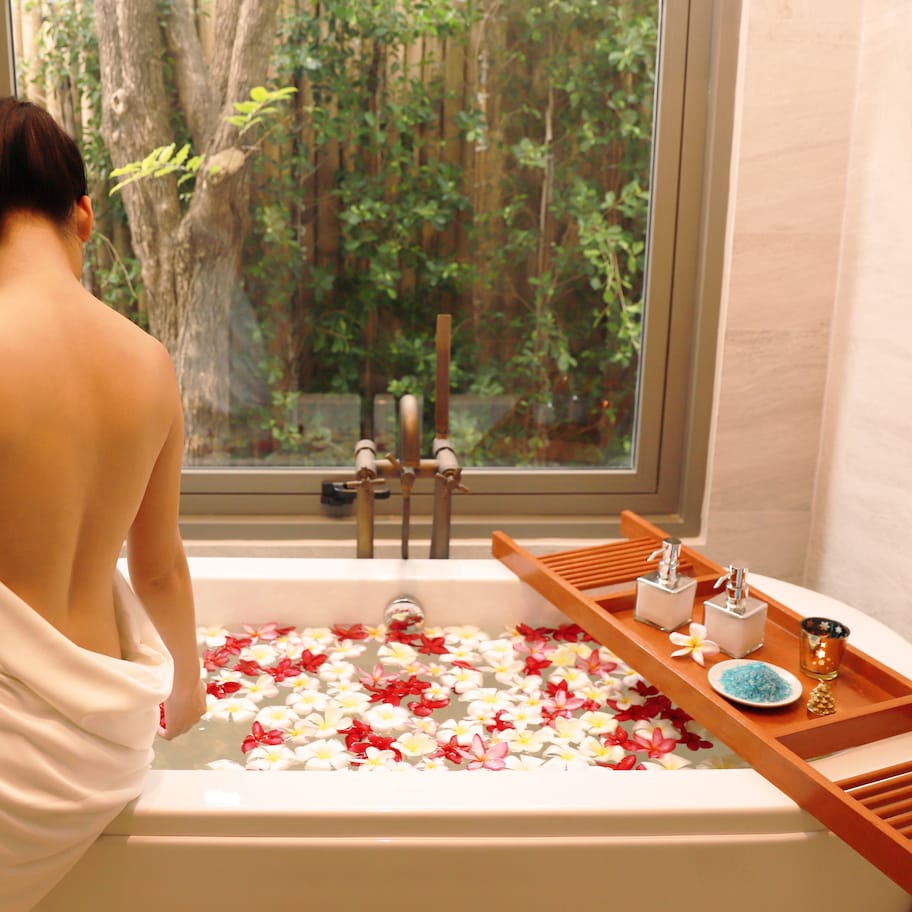 A bathtub filled with flower petals, a wooden tray with toiletries, and a window with a forest view.