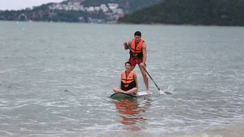 A person in a life jacket standing on a paddleboard with another person sitting on it.
