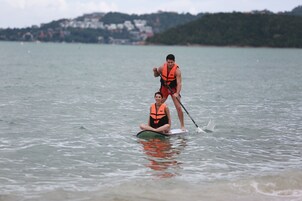 A person in a life jacket standing on a paddleboard with another person sitting on it.