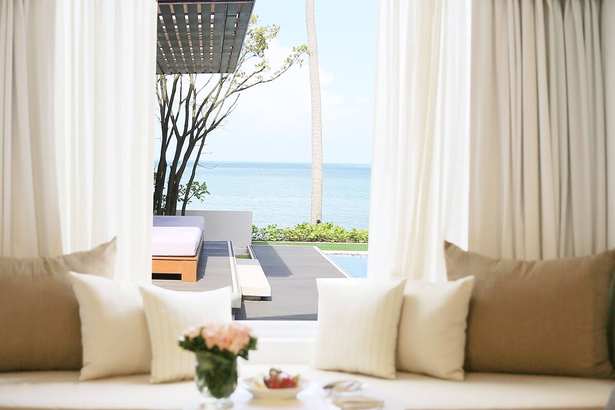 A room with a view of the sea, a sofa with cushions, and a small table with a vase.