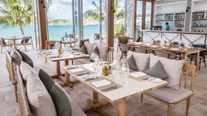 Un restaurant situé en bord de mer, doté de nombreuses tables et chaises, de grandes fenêtres offrant une vue sur l’océan, et d’un bar visible au fond.