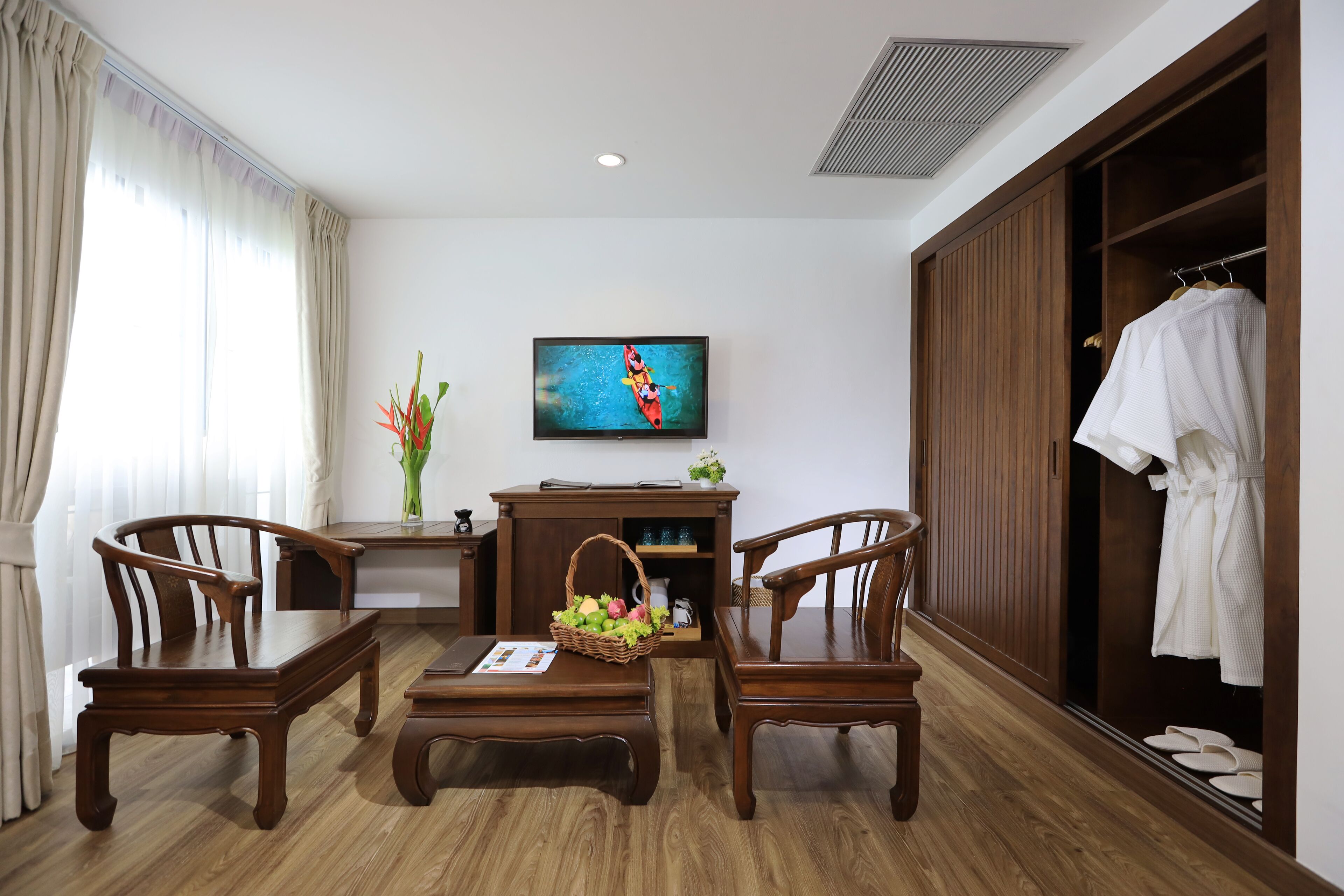 A hotel room with a wooden floor, a TV mounted on the wall, a wooden wardrobe, and a seating area with two wooden chairs and a table.