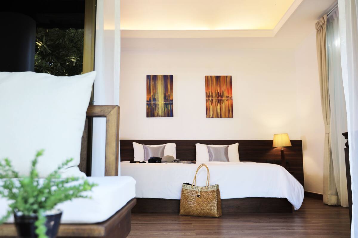 A hotel room with a large bed, two wall-mounted paintings, a bedside lamp, and a wicker handbag on the floor.