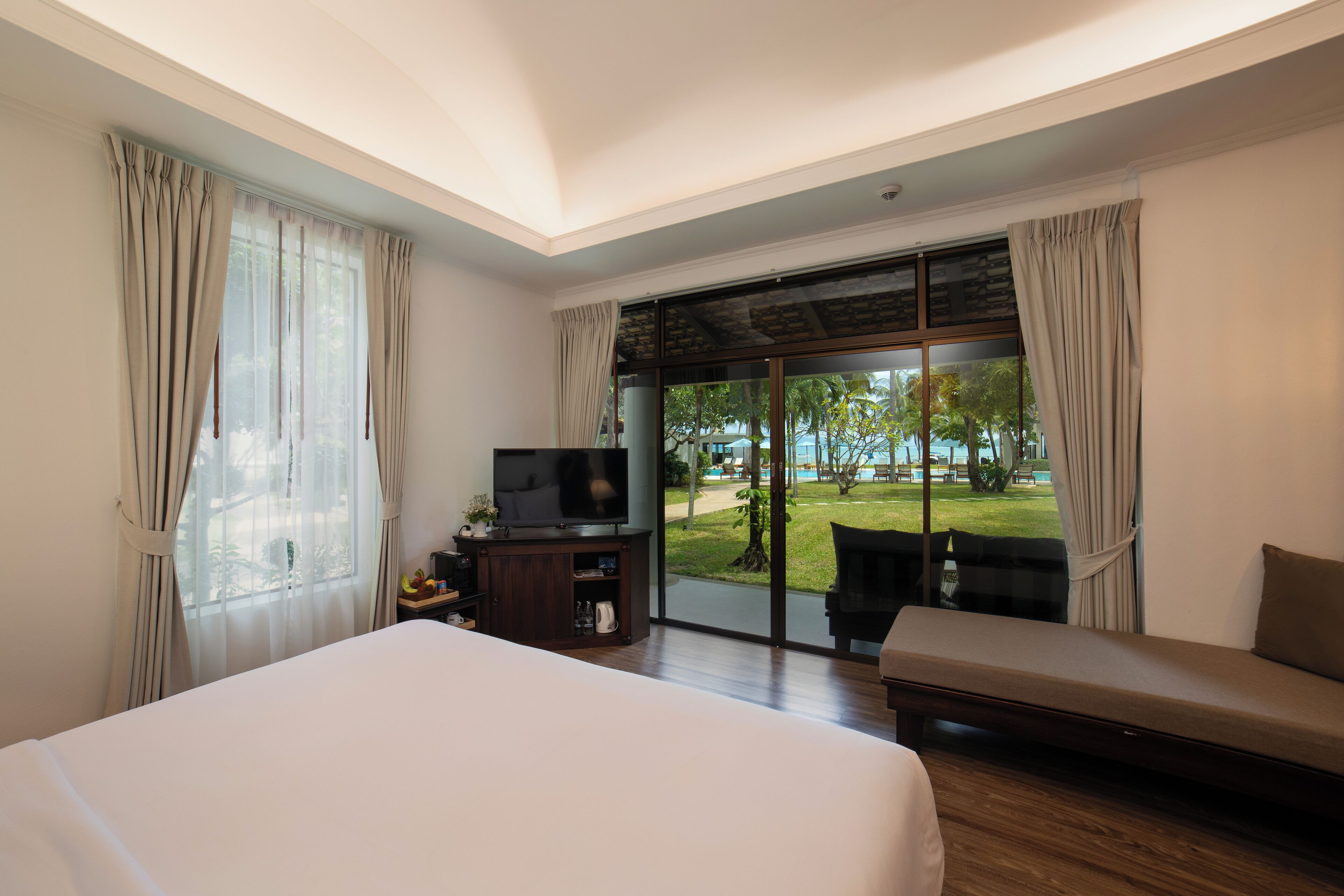 Camera d'albergo con un letto grande, una TV, un tavolino con una pianta e vista su giardino e piscina.