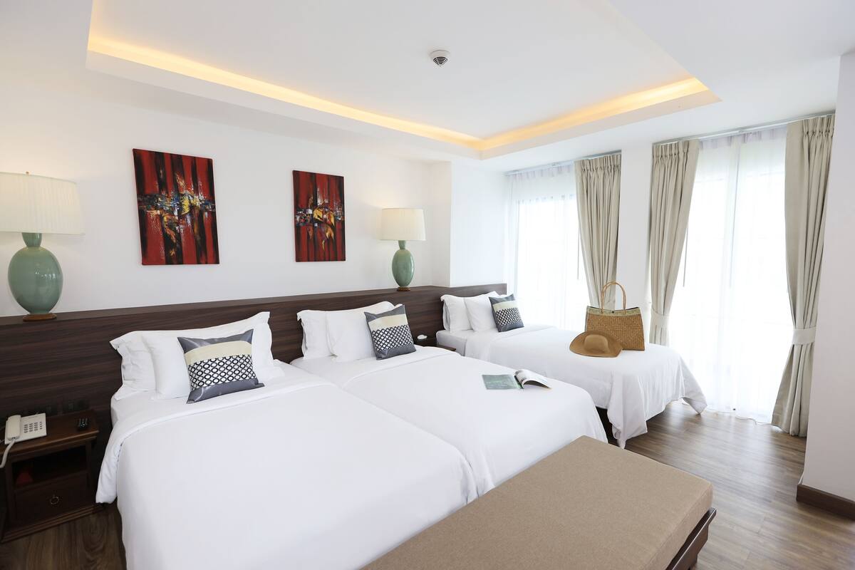 A hotel room with two beds, a bedside table with a lamp, a wall-mounted phone, and a window with curtains.