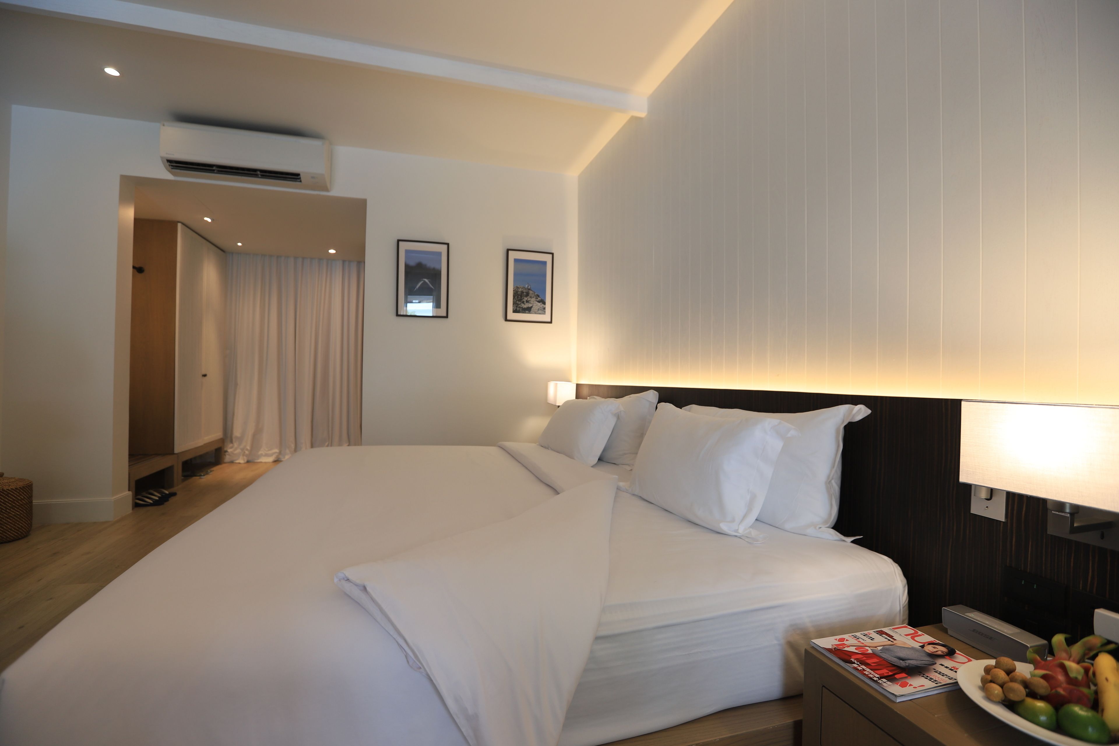 A hotel room with a large bed, a bedside table with a magazine and fruit, and a wall-mounted lamp.
