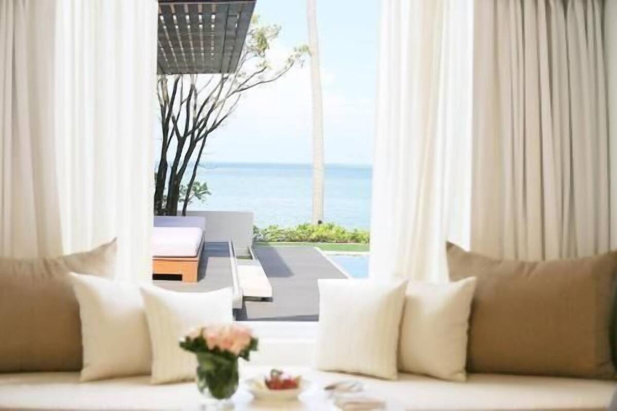 A room with a view of the sea, a sofa with cushions, and a small table with a vase.