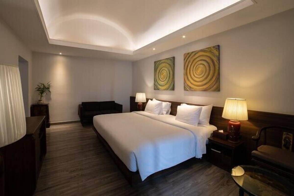 A modern hotel room with a large bed, two bedside tables with lamps, a dark sofa, and abstract wall art.