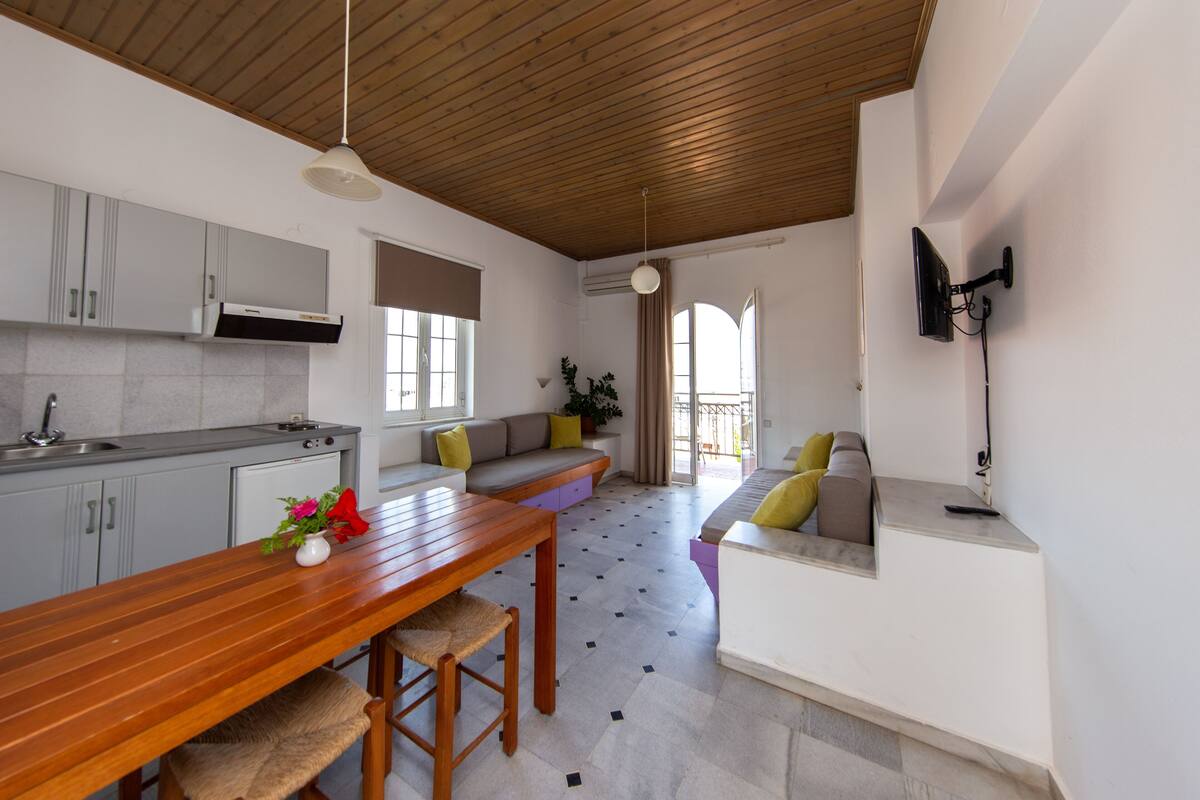 A modern kitchen with a wooden ceiling, a dining table with chairs, a sofa with cushions, and a TV mounted on the wall.