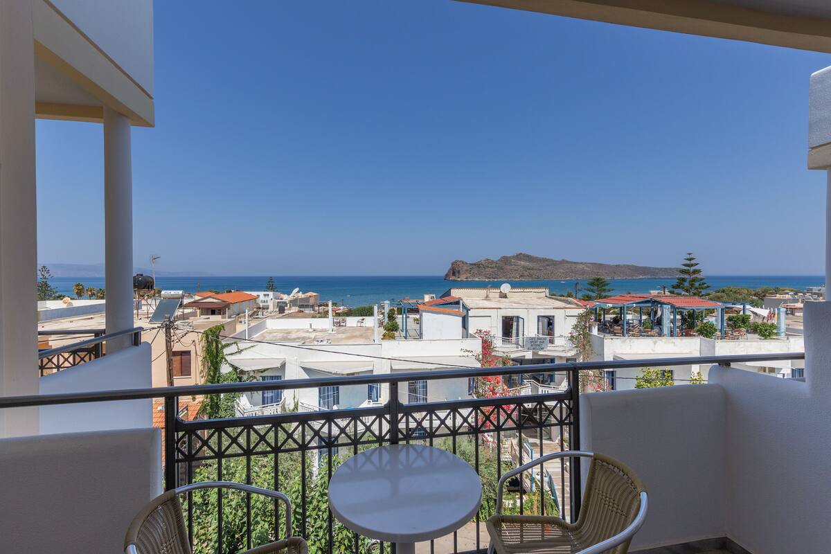 A balcony with a table and chairs, overlooking a coastal town with buildings and a mountain in the distance.