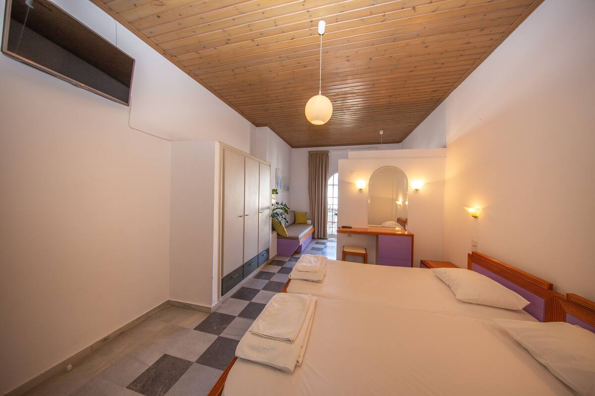 A hotel room with a checkered floor, a bed with white linens, a wooden ceiling, and a small desk with a mirror.